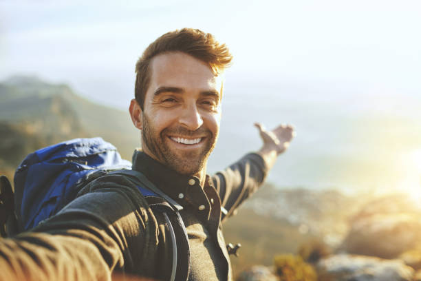 A selfie of John. He is outside with the sun behind him. John has a beard and short brown hair. He is smiling and holding his hand out reached towards the sun. He also has a hiking pack on his back.